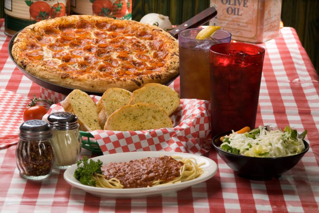 Pizza, garlic bread, pasta, salad, salt, pepper, and drinks set on a red and white table cloth.